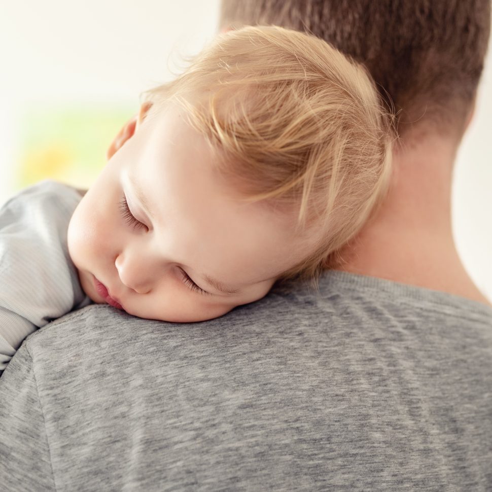 Portrait of cute adorable blond caucasian toddler boy sleeping on fathers shoulder indoors. Sweet little child feeling safety and care on daddys hand. Single father concept.